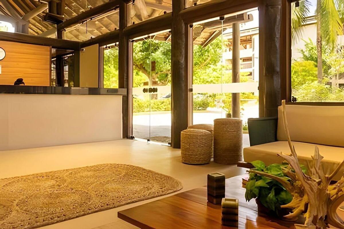 Ein modernes Interieur mit einem Holzerrezeptionstresen, einem großen Fenster mit Blick auf Grünflächen, einer Sitzecke mit Sofa und Poufs sowie einem Couchtisch aus Holz mit einer Pflanze.