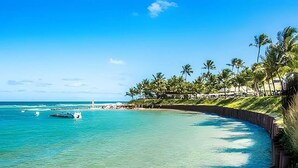 A coastal view with clear turquoise waters, a lighthouse, and palm trees. - Condomínio Nannai Residence (Ipojuca)