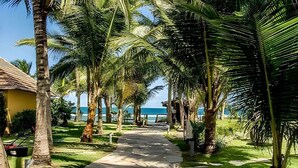 A pathway lined with palm trees leading to a beach, with a small building on the left. - Condomínio Nannai Residence (Ipojuca)