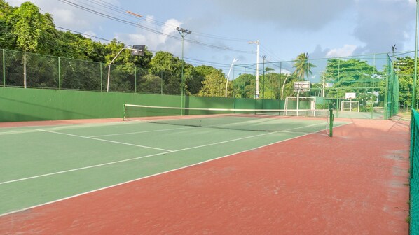 A tennis court with a net, surrounded by a green fence and trees. - Condomínio Nannai Residence (Ipojuca)