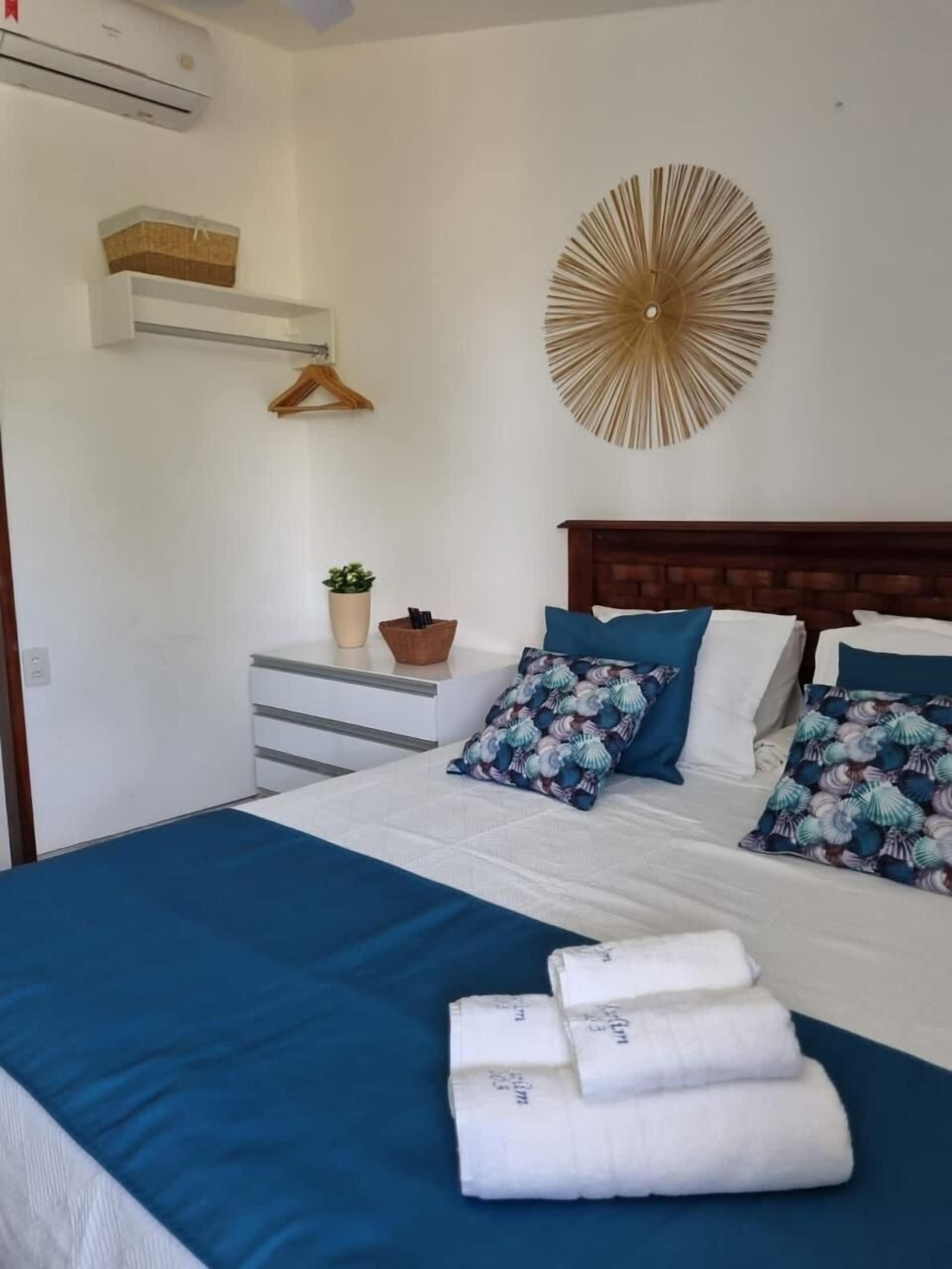 A neatly made bed with a blue bedspread and white pillows, a decorative wall piece, a small potted plant, and a wooden hanger on a white dresser.