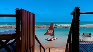 A beach view from a wooden deck with a sailboat in the water. - Condomínio Nannai Residence (Ipojuca)