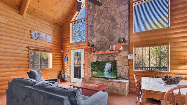 A log cabin interior with a stone fireplace, wooden walls, a sofa, a dining table, and a TV. - The Family Bear Cabin at Mt. Lemmon Hotel (Mount Lemmon)