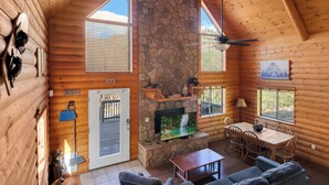 A log cabin interior with a stone fireplace, wooden walls, a dining area, and a television. - The Family Bear Cabin at Mt. Lemmon Hotel (Mount Lemmon)