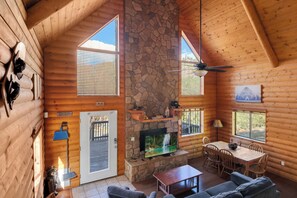 Interior de uma cabana de toras com lareira de pedra, paredes de madeira, área de jantar e uma televisão.