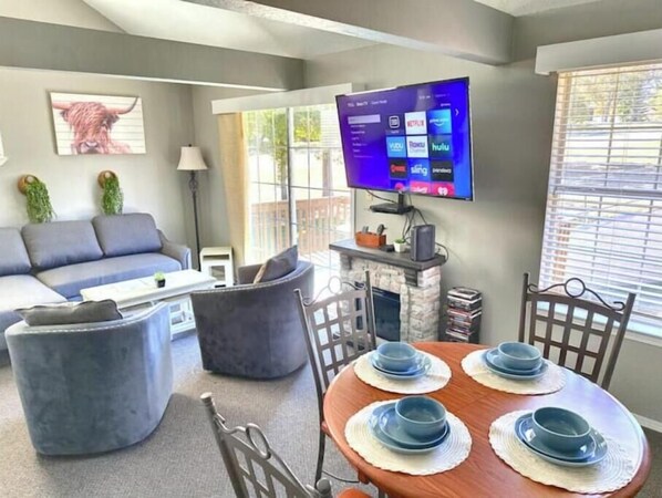 A modern living room with a sofa, ottoman, and a dining table set with blue plates. A flat-screen TV is mounted on the wall, and there is a fireplace.