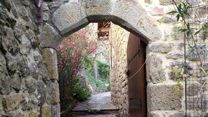 Un chemin de pierre mène à une porte en arc, bordée de murs en pierre et agrémentée d’un pot de fleurs.