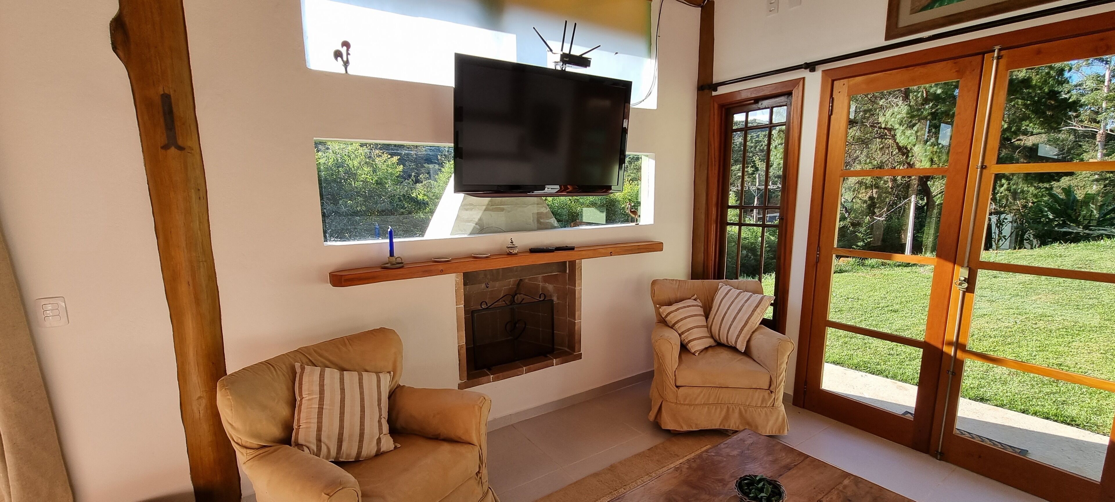 A living room with a TV mounted above a fireplace, two armchairs, and a large sliding glass door leading to a garden.