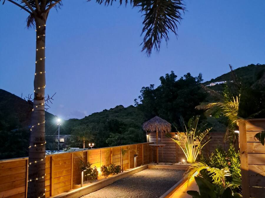 Un espacio exterior bien iluminado con una terraza de madera, palmeras y una estructura con techo de paja.