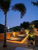 Un espacio exterior bien iluminado con una terraza de madera, palmeras y una estructura con techo de paja.