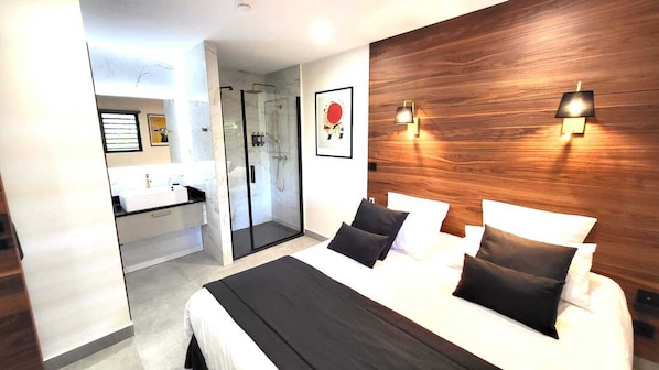A modern hotel room with a large bed, a glass-enclosed shower, a wooden accent wall, and a framed picture on the wall. - The Forest Suites at Anse Marcel | Boutique Hotel & Restaurant (Anse Marcel)