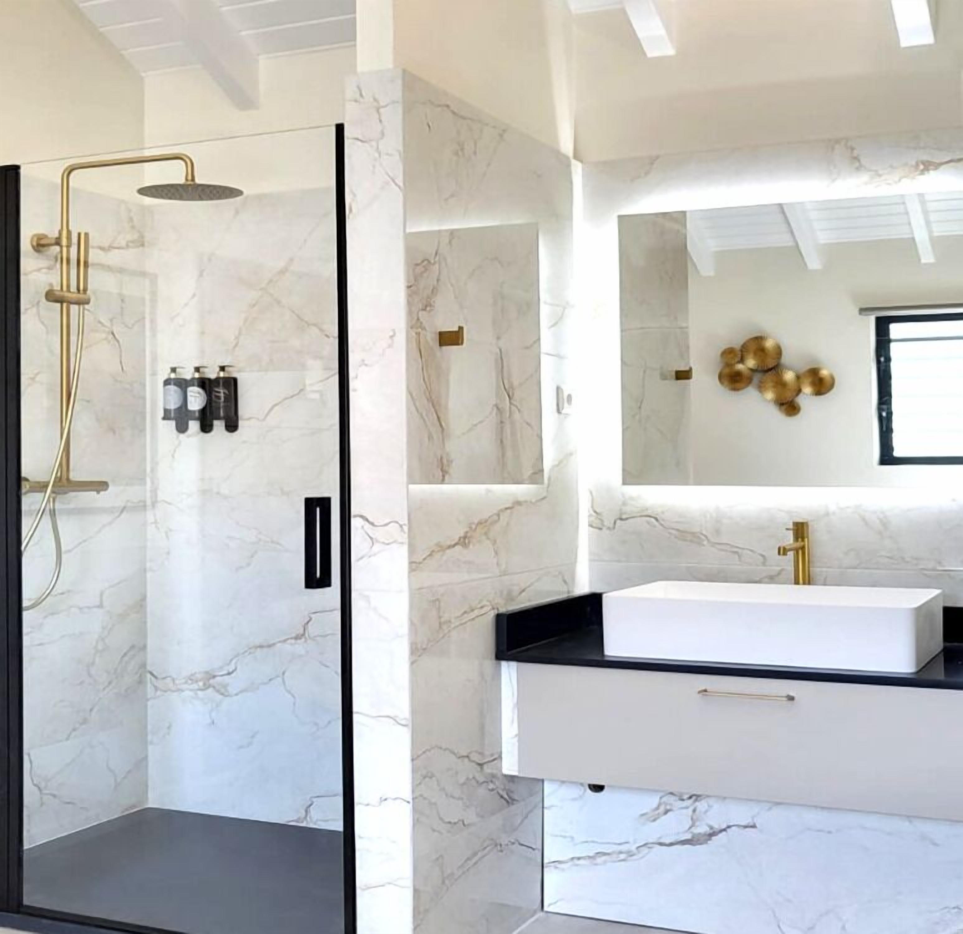 A modern bathroom with a glass-enclosed shower, marble walls, and a white vanity with a gold faucet.