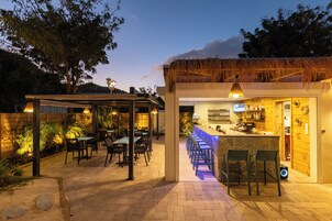 Un patio al aire libre con mesas y sillas, una zona de bar con taburetes y una estructura de techo de paja.