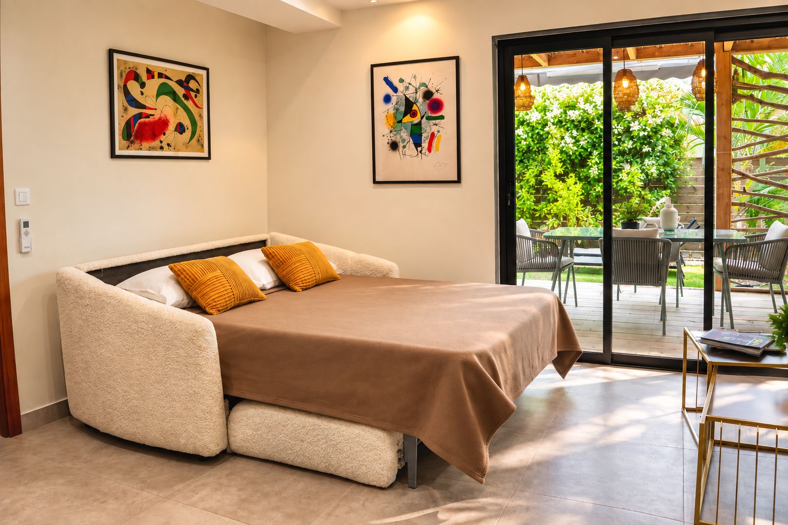Een moderne slaapkamer met een slaapbank, twee ingelijste abstracte schilderijen en uitzicht op een terras met stoelen en begroeiing.