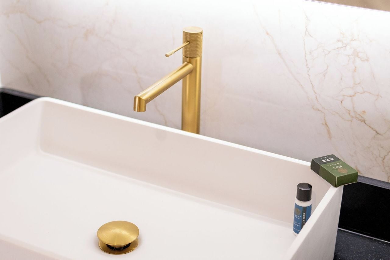 A bathroom with a gold faucet, a white sink, and a bottle of lotion.
