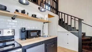 A compact kitchen with a built-in oven, microwave, and sink. There is a wooden shelf above the counter holding dishes and a coffee maker. A staircase with a metal railing leads to an upper level. - Chic Cabin! Less than 10 minutes from Downtown Siloam Springs (Gentry)
