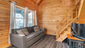 Interior de una cabaña de madera con sofá, televisión y una escalera.