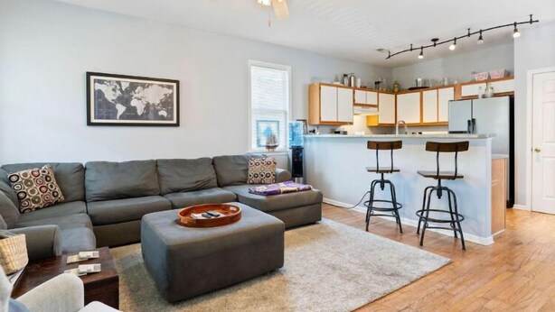 Un salon moderne avec un canapé gris, un pouf et une table basse. On y trouve également un coin cuisine avec un tabouret de bar et un réfrigérateur.