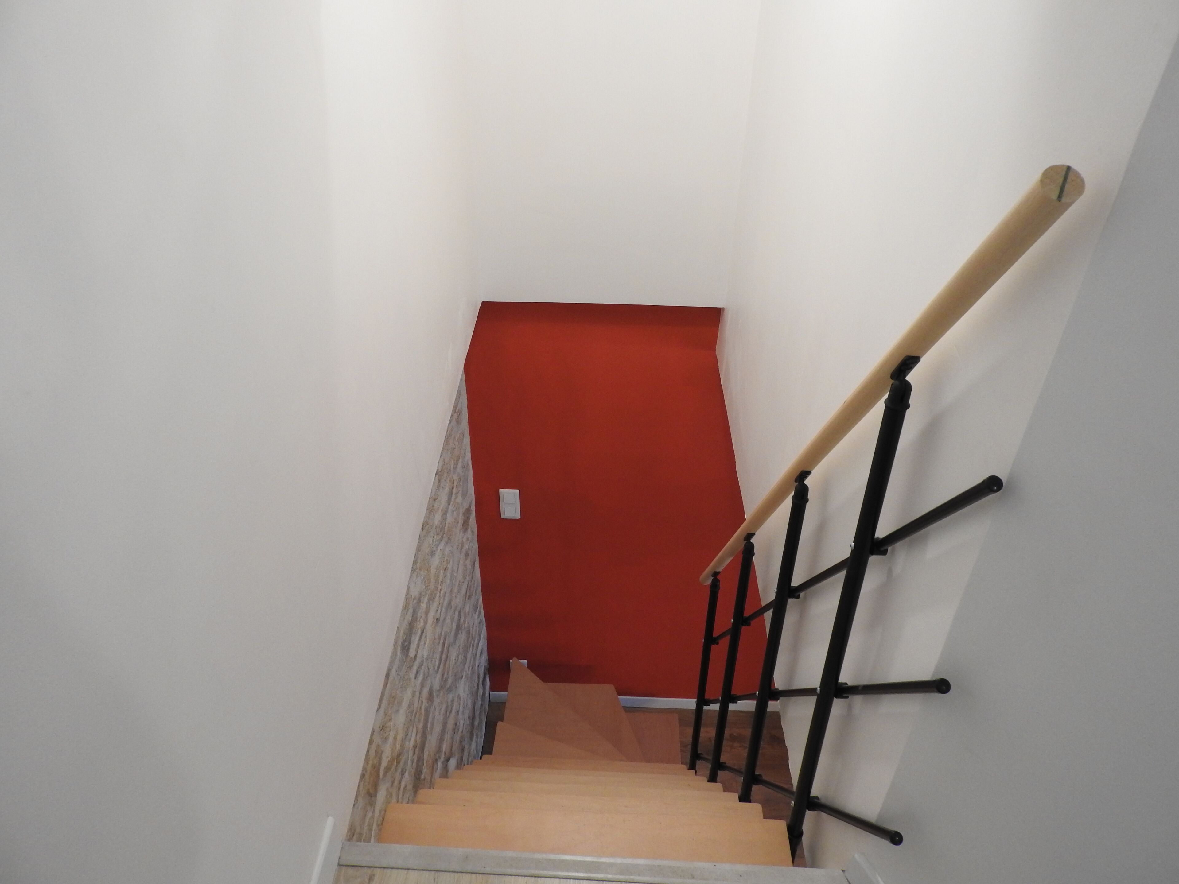 A staircase with a red wall at the top, a wooden handrail, and a white wall on the left.