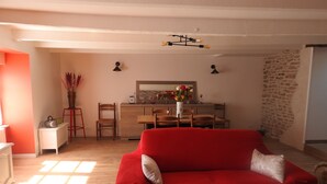 Ein gemütliches Wohnzimmer mit rotem Sofa, Esstisch aus Holz und Stühlen. Es gibt eine Küchenzeile mit Holzschränken und einer Pendelleuchte.