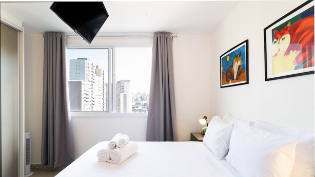 Habitación de hotel con cama, ventana con cortinas, televisor de pared y dos cuadros enmarcados.