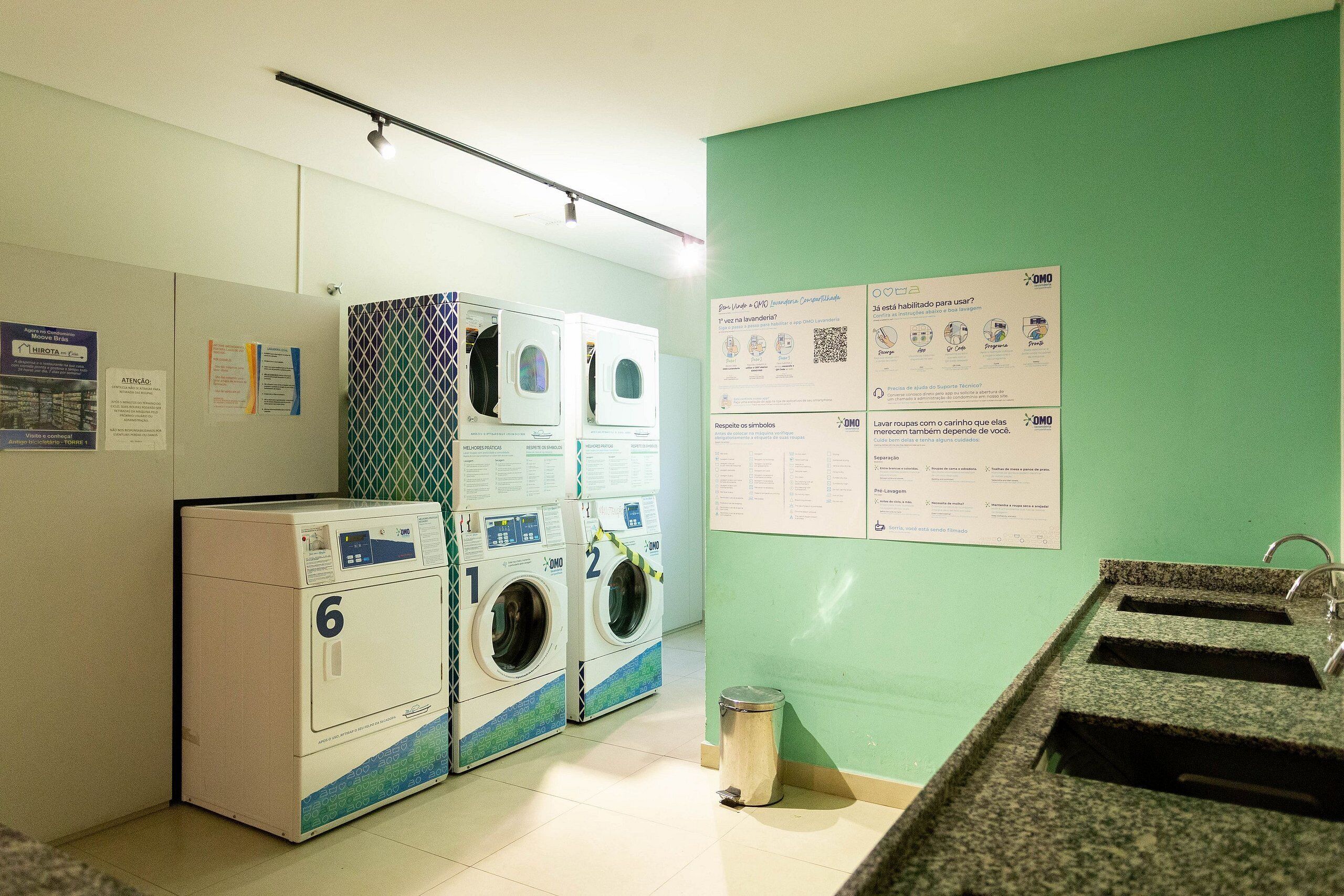 A laundry area with multiple washing machines, a trash bin, and informational posters on the wall.