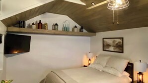 Un dormitorio con techo inclinado, una cama, lámparas de noche, un televisor montado y un estante con diversos objetos.