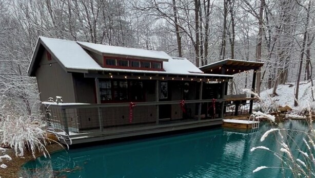 Escena invernal con un edificio junto a una piscina azul cristalina, rodeado de tierra y árboles cubiertos de nieve.