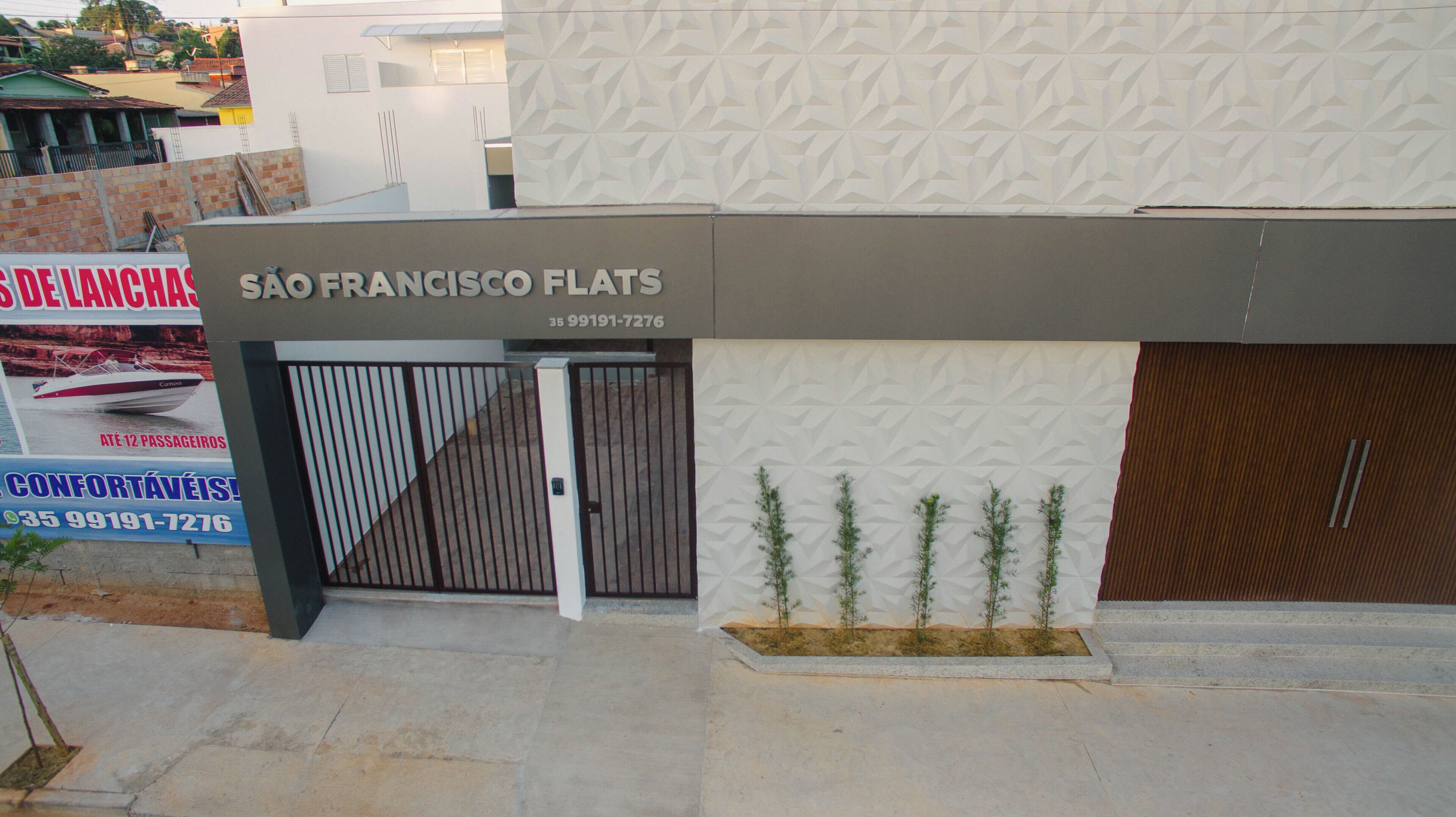 Un immeuble avec une enseigne indiquant « SÃO FRANCISCO FLATS » et un numéro de téléphone. L’entrée est sécurisée par un portail en métal et un petit jardinier avec des plantes est placé devant.