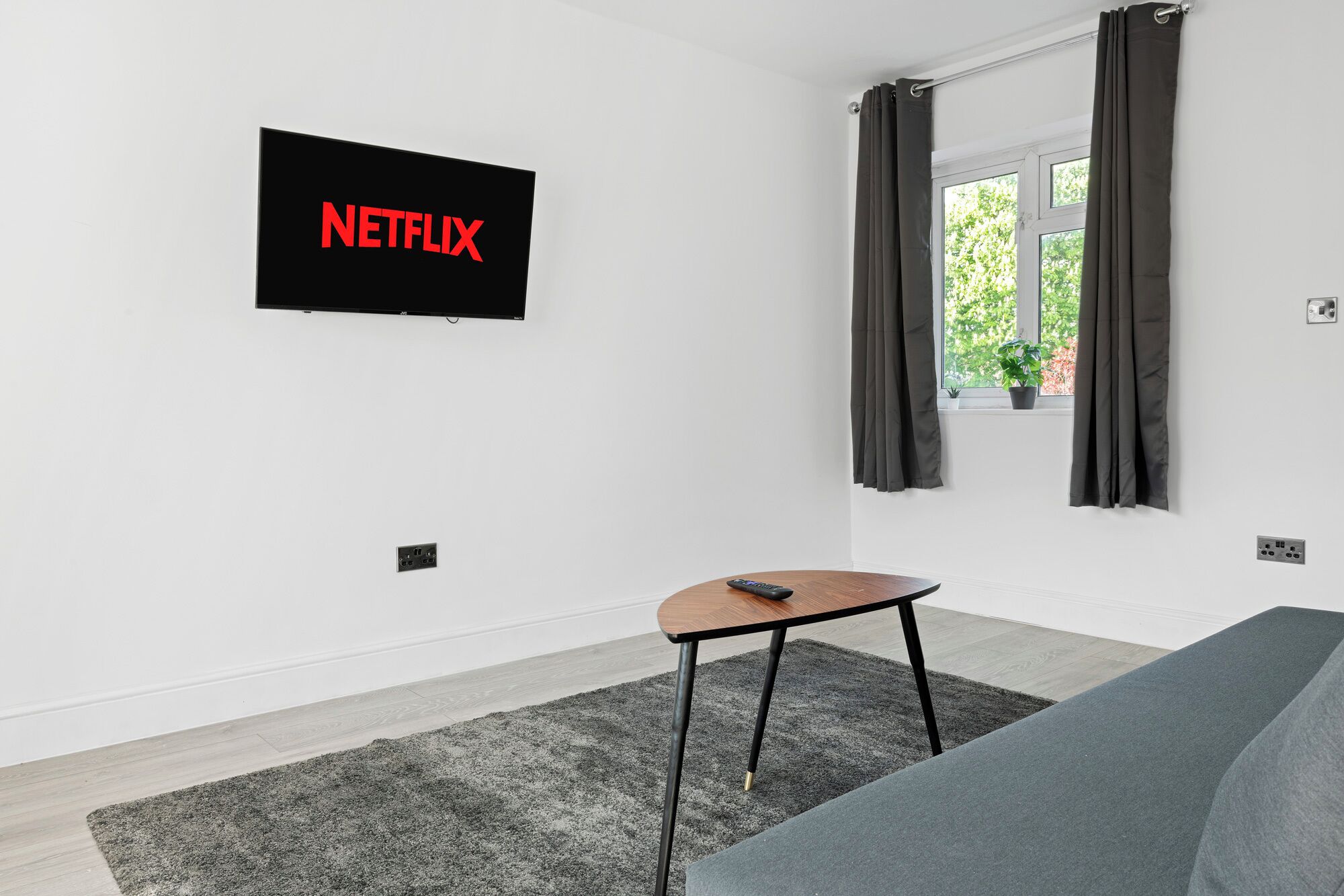 A modern living room with a flat-screen TV mounted on the wall displaying the Netflix logo, a small wooden table, a grey sofa, and a window with dark curtains.