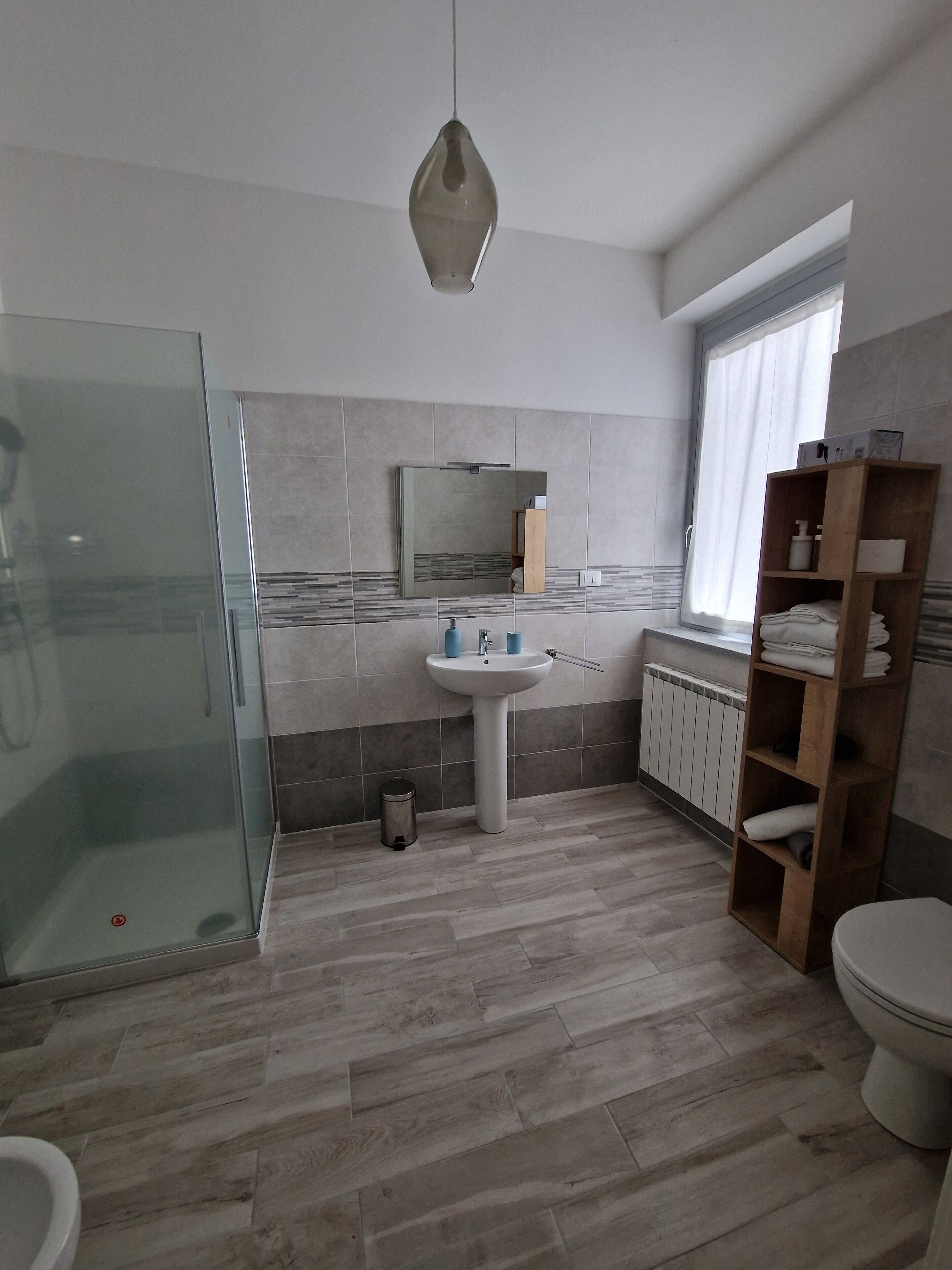 A modern bathroom with a glass shower enclosure, a pedestal sink, a wooden shelf with towels, and a window with curtains.