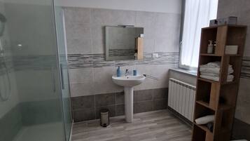 A modern bathroom with a glass shower enclosure, a pedestal sink, a wooden shelf with towels, and a window with curtains.