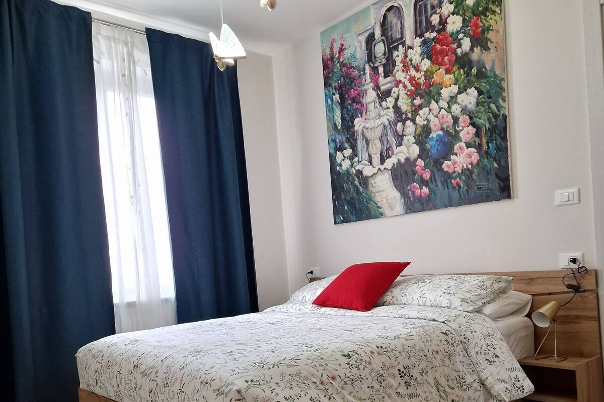 A bedroom with a wooden bed, floral bedding, a red pillow, a nightstand, a lamp, and a large floral painting on the wall.