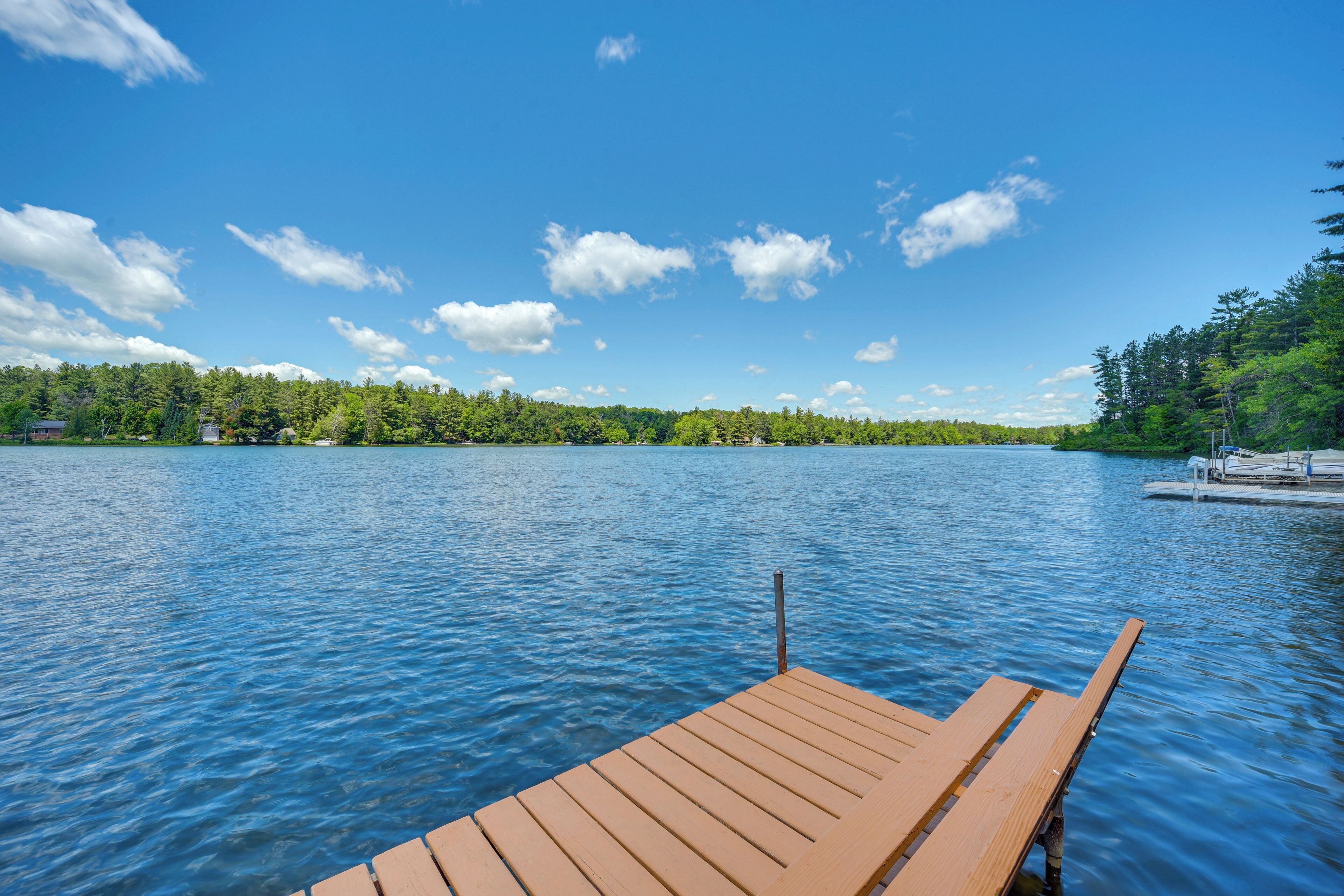 Eine Holzsteganlage, die in einen ruhigen See hineinreicht, mit blauem Himmel und bewaldeter Uferlinie.