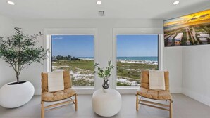 A room with two chairs facing a large window with a view of the beach and ocean. There is a potted plant and a vase on a round table in the center. - Shangri-La | Bean Point | Rooftop Pool | Golf Cart (Anna Maria)