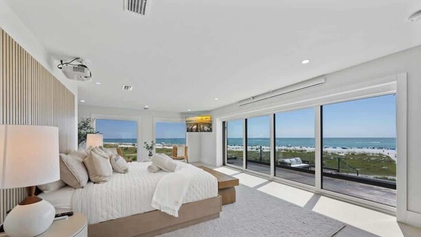 A bedroom with a large bed, a nightstand, a lamp, and a view of the beach. - Shangri-La | Bean Point | Rooftop Pool | Golf Cart (Anna Maria)