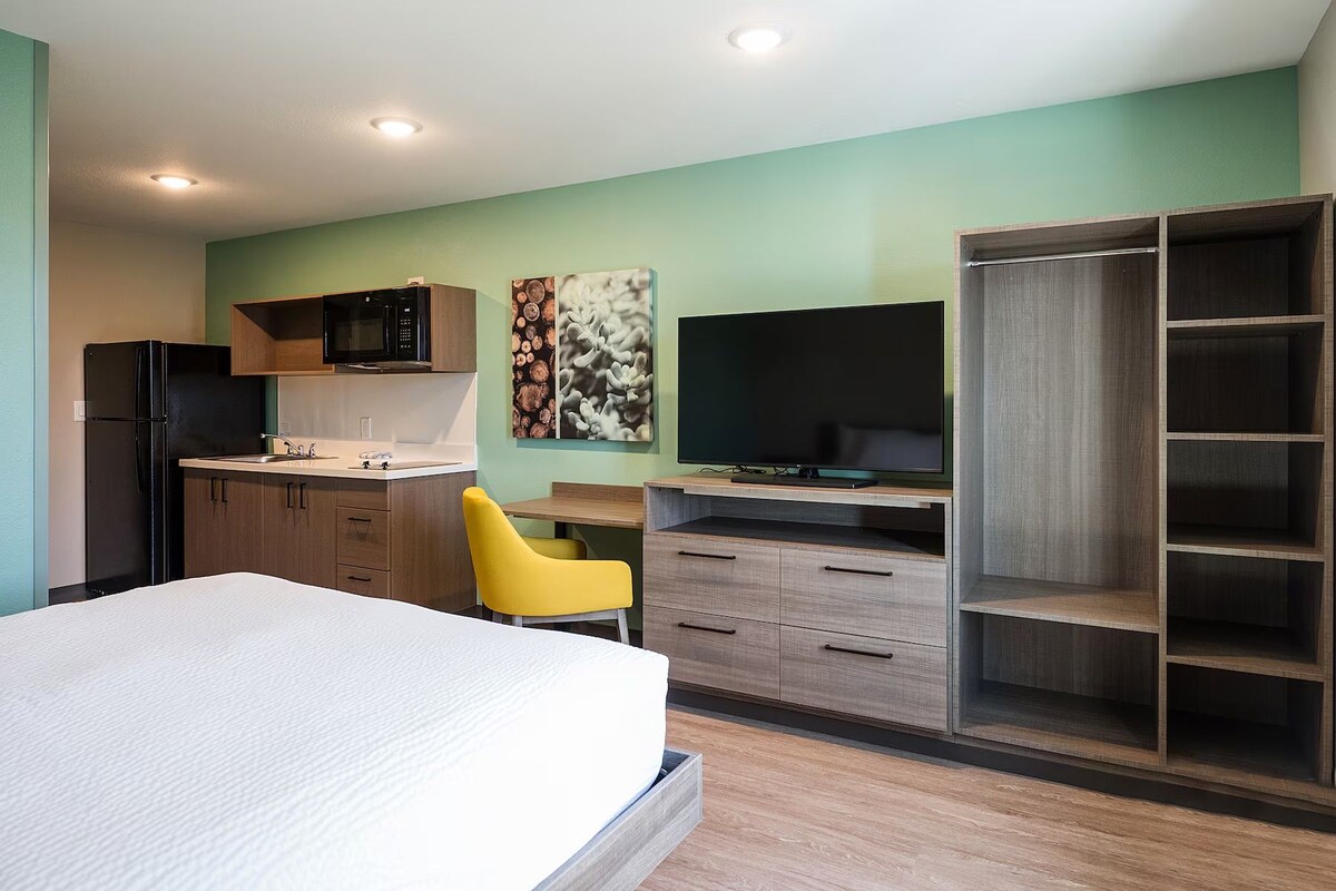 Habitación compacta con cama, escritorio, silla, televisor, armario y kitchenette.