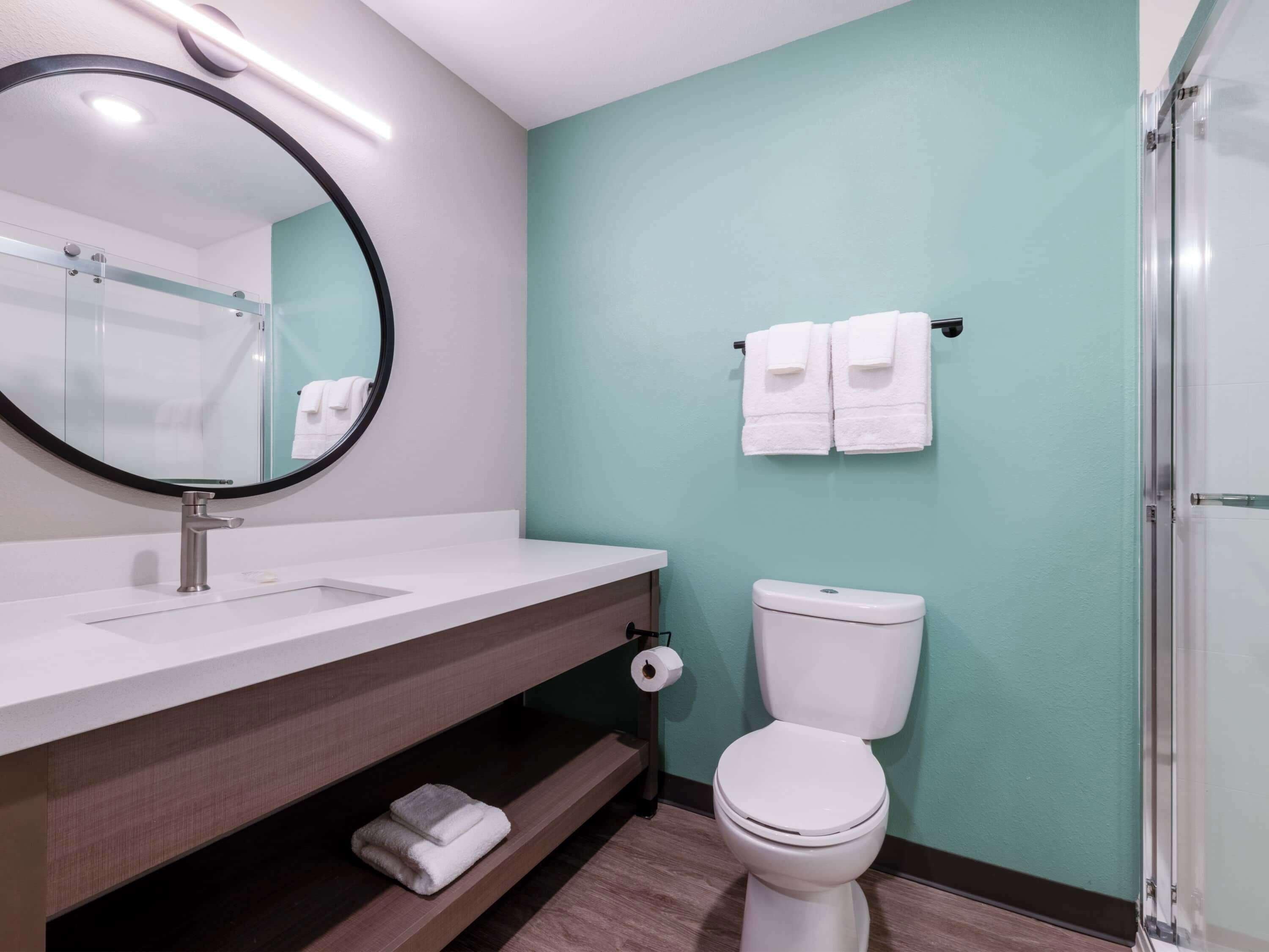A modern bathroom with a white sink, a round mirror, a toilet, and a glass shower enclosure.