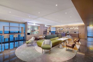 Un moderno vestíbulo de hotel con un sofá verde, sillones blancos, una barra de café y vistas a la ciudad.