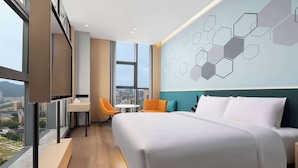 Une chambre d’hôtel moderne dotée d’un grand lit, d’un bureau et d’une chaise, offrant une vue sur la ville et présentant un motif géométrique sur le mur.