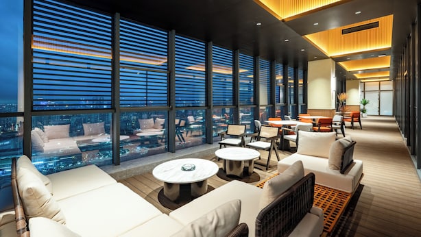 Un salon moderne situé sur le toit, doté de grandes fenêtres, de sièges confortables et offrant une vue sur la ville.