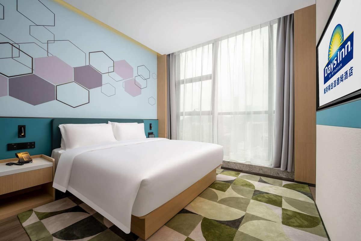 Habitación de hotel con una cama grande, una mesita de noche con teléfono, una pared con patrones geométricos, un ventanal con cortinas y un logotipo en la pared.