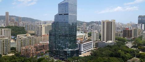 A high-rise building with a modern glass facade, surrounded by other residential and commercial buildings, and a park area with trees.