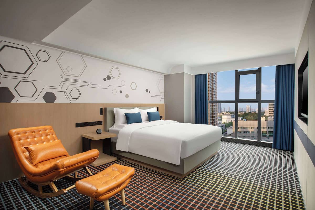 Habitación de hotel moderna con una cama grande, un sillón de cuero y vistas a la ciudad a través de un ventanal.