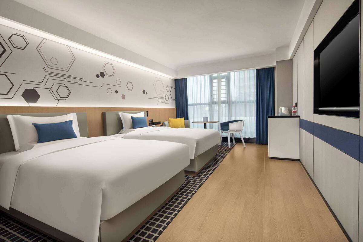Una habitación de hotel con dos camas, un escritorio, un televisor y un ventanal grande con cortinas.