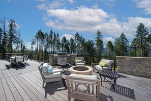 Relax on the deck with a fire pit, lounge chairs, and dining table, surrounded by tall trees and a serene view.