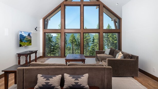 A spacious living room with a large window offering a mountain view, a cozy sofa set, and a wooden coffee table. - Great Northern Mountain Escape (Whitefish)