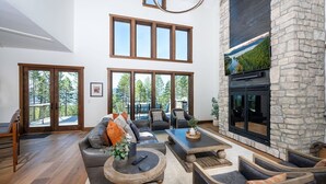 A spacious living room with a stone fireplace, large windows, and a view of the outdoors. - Great Northern Mountain Escape (Whitefish)