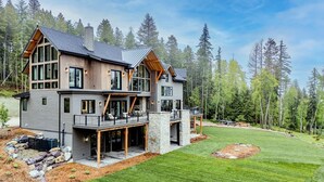 A modern two-story house with a large deck, surrounded by a well-maintained lawn and dense forest. - Great Northern Mountain Escape (Whitefish)