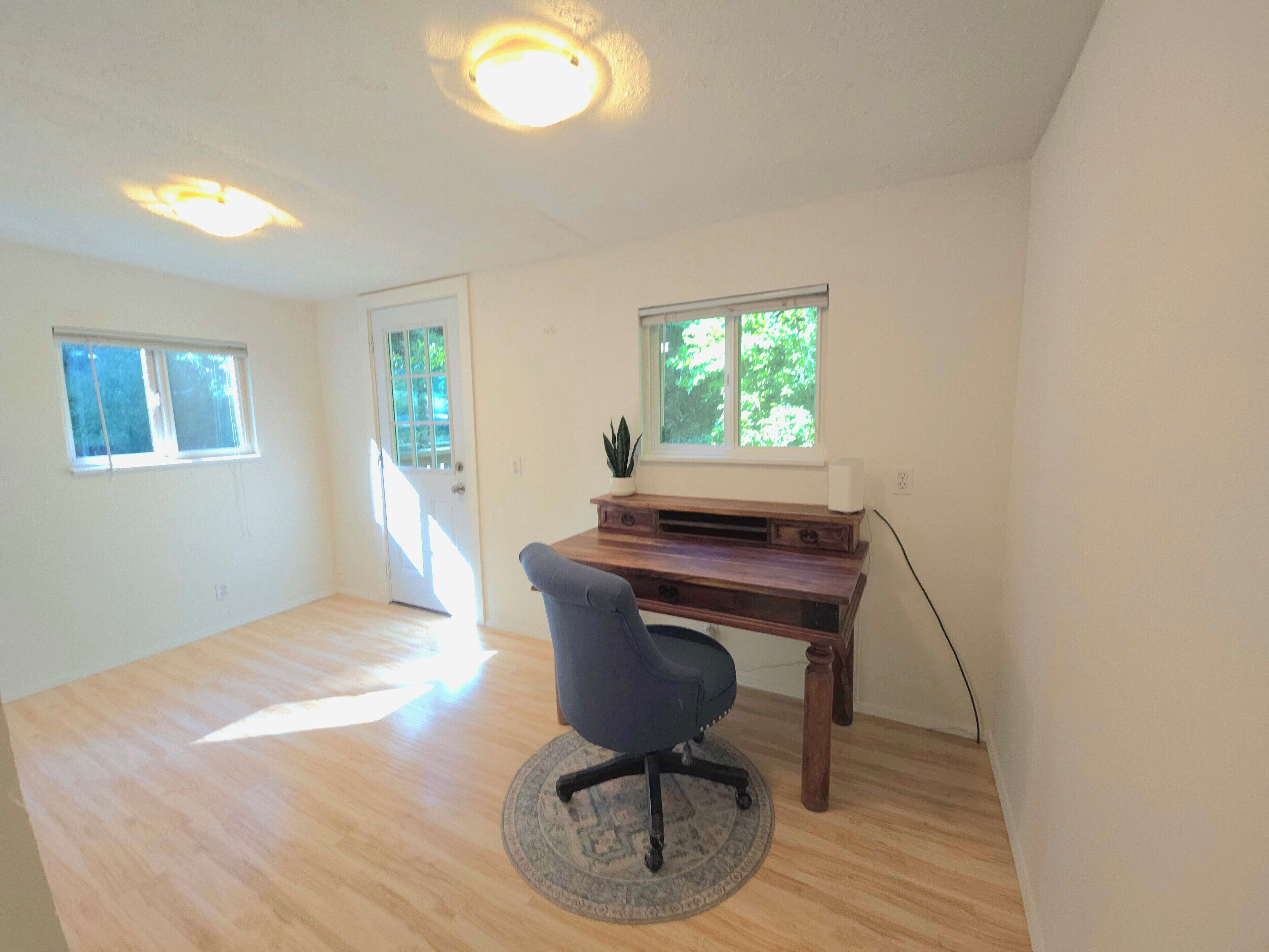 Ein geräumiges Zimmer mit Holzboden, Schreibtisch mit Computer, Stuhl und zwei Fenstern.